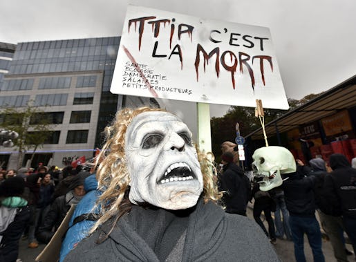 A protestor with a mask demonstrates against the free trade agreement TTIP (Transatlantic Trade and Investment Partnership) during an EU summit in Brussels, Belgium on Thursday, Oct. 15, 2015. His banner reads "TTIP is the dead". European Union heads of state meet to discuss, among other issues, the current migration crisis. (AP Photo/Martin Meissner)