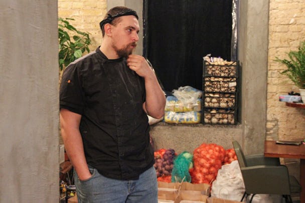 Kocken Pavlo Khrobust, i svart kockjacka och svart bandana runt huvudet, lutar sig mot en pelare medan han drar fingrarna genom skägget. I bakgrunden står nätsäckar med grönsaker.