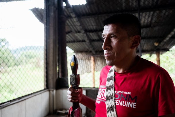 Urfolksledaren Marcelino Noscue, iklädd röd t-tröja och ett virkat band över bröstet, blickar ut i fjärran. I ena handen håller han en snidad trästav.