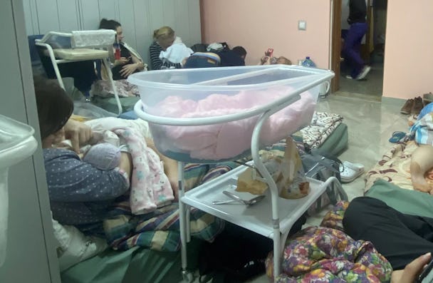 En barnsäng i plast med rosa täcke står på golvet i sjukhusets skyddsrum i Kiev. På madrasser längs väggarna sitter mammor med bebisar i sina armar.