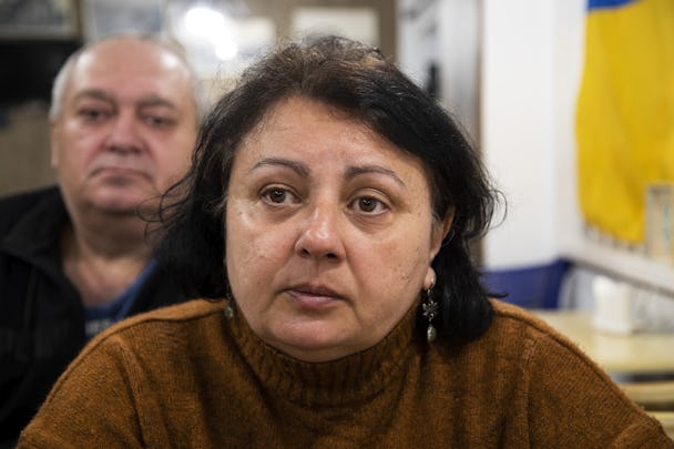 En mörkhårig kvinna med brun tröja tittar strax till vänster om kameran. Hon har silverörhängen med gröna stenar i. Till höger i bild hänger en ukrainsk flagga på en vägg. Längre bak syns kvinnans make oskarpt.