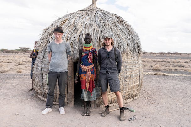 Två män står på vardera sidan av en kvinna från Turkanafolket, framför en hydda byggd av löv och grenar. Mannen till vänster bär grå byxor, grå t-tröja och svart hatt. Mannen till höger bär grå shorts, grå skjorta och keps. Kvinnan i mitten har färglada kläder och flera tjocka halsband runt halsen. Landskapet bakom hyddan är kargt.