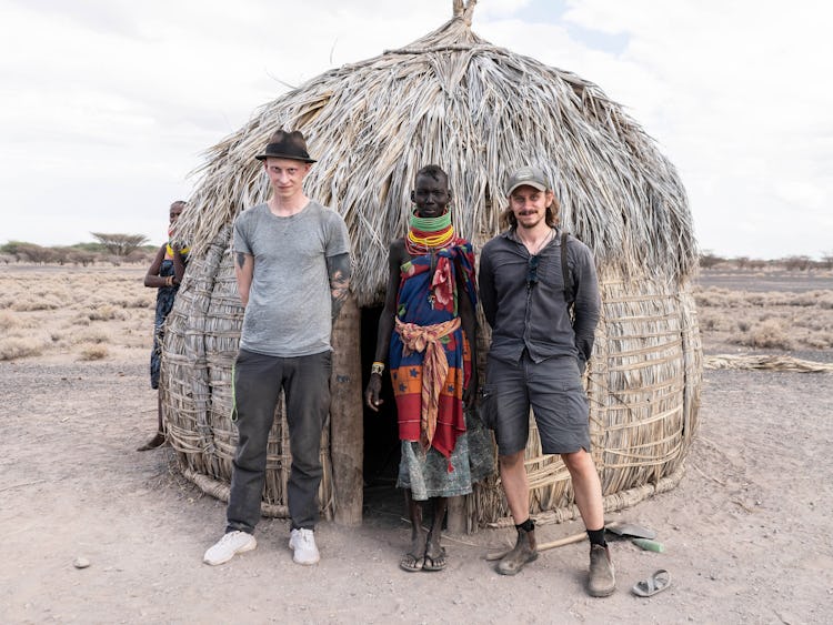 Två män står på vardera sidan av en kvinna från Turkanafolket, framför en hydda byggd av löv och grenar. Mannen till vänster bär grå byxor, grå t-tröja och svart hatt. Mannen till höger bär grå shorts, grå skjorta och keps. Kvinnan i mitten har färglada kläder och flera tjocka halsband runt halsen. Landskapet bakom hyddan är kargt.