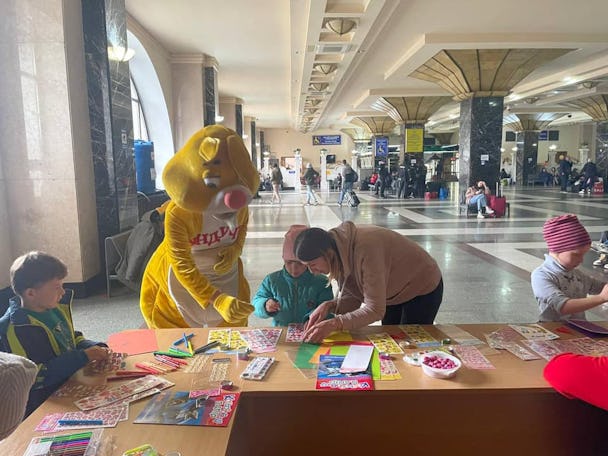 Tre barn sitter vid ett bord fyllt av vykort och pysselmaterial i vänthallen på en tågstation. En blond kvinna böjer sig över barnet i mitten. Till vänster om barnet står en person utklädd till en gul påskhare.