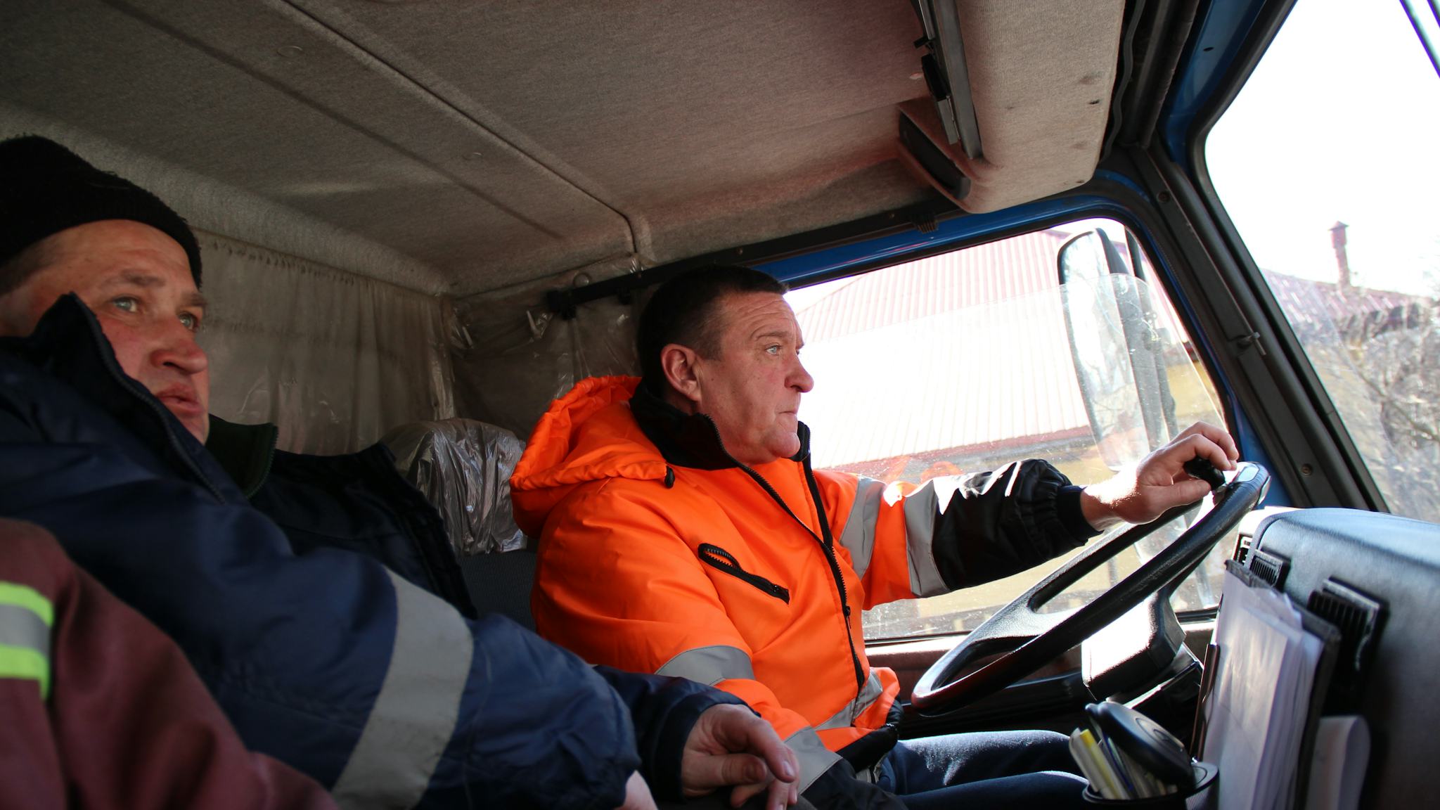 En sopåkare i kortklippt mörkt hår och orange varseljacka sitter bakom ratten tille n sopbil. Till vänster syns en kollega i mörkblå jacka och svart mössa.