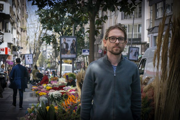 En man i 40-årsåldern med svarta glasögon och axellångt hår står på en gata i Istanbul. I bakgrunden syns en blomsterförsäljares stånd.