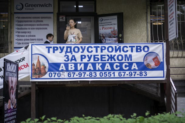 En vit skylt med blå kirgizisk text gör reklam för jobb i Ryssland. Skylten sitter uppspänd på en veranda. Ovanför skylten står en ung kvinna och tittar på en mobilskärm.