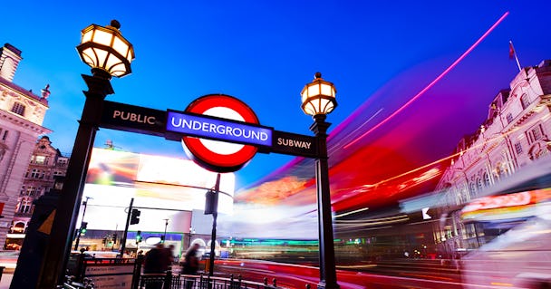 Brittisk tunnelbaneskylt vid Piccadilly Circus i London fotograferad mot natthimmel. Ljus från trafik susar förbi.