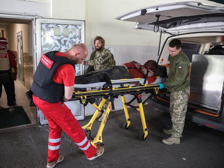 En ambulanssjukvårdare i röda kläder samt två män i militärkläder flyttar en bår från en ambulans för att köra in den genom en sjukhusentré. På båren ligger en skadad man med ansiktet dolt i skugga.