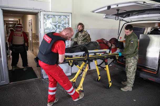 En ambulanssjukvårdare i röda kläder samt två män i militärkläder flyttar en bår från en ambulans för att köra in den genom en sjukhusentré. På båren ligger en skadad man med ansiktet dolt i skugga.