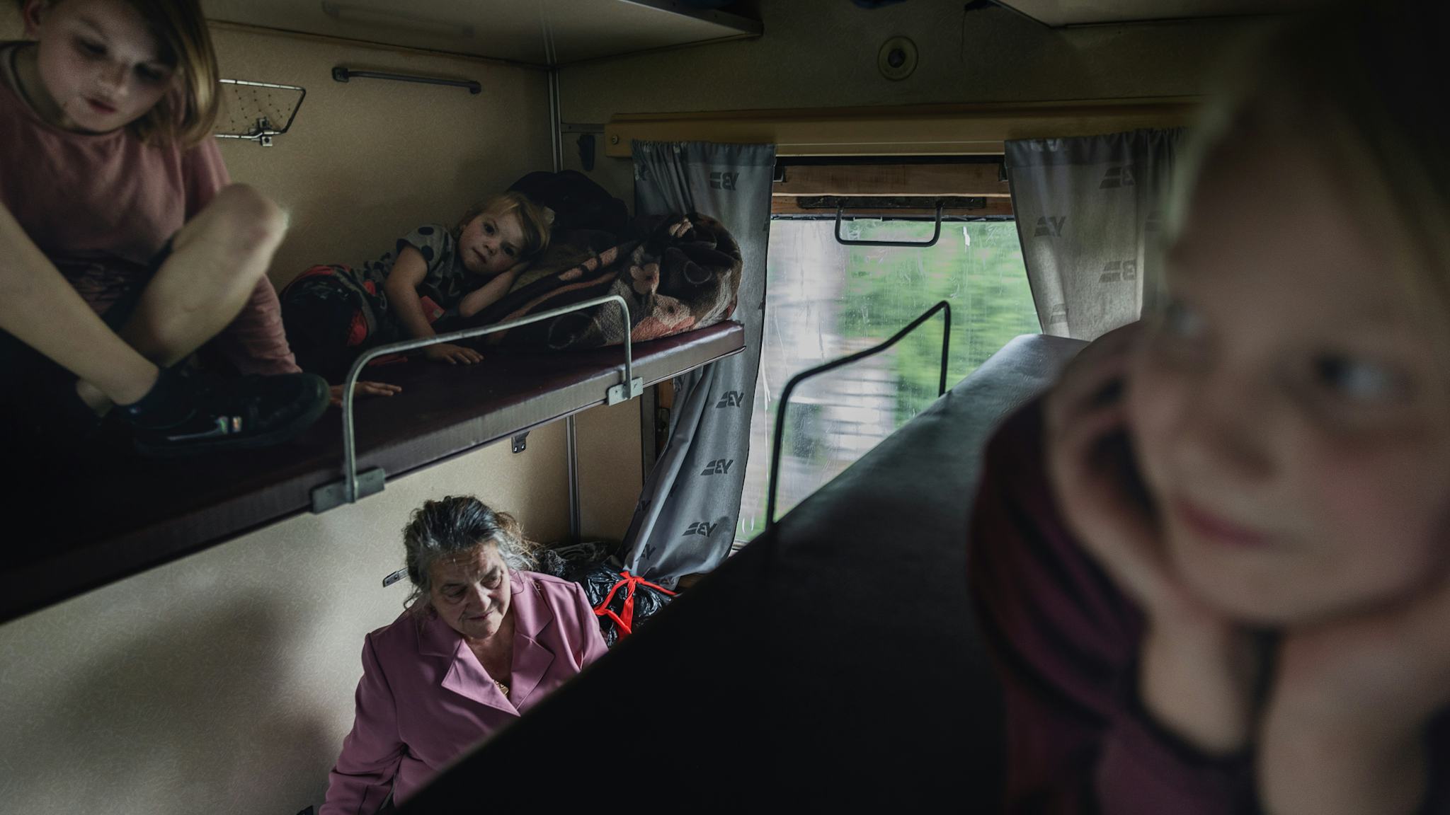 Tre blonda barn sitter på varsin överslaf i en tågkupé. På britsen nedtill syns en äldre gråhårig kvinna i Rosa kappa.
