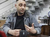 En mörkhårig man med skäggstubb, iklädd jeansjacka, sitter vid ett bord med en kaffekopp framför sig.