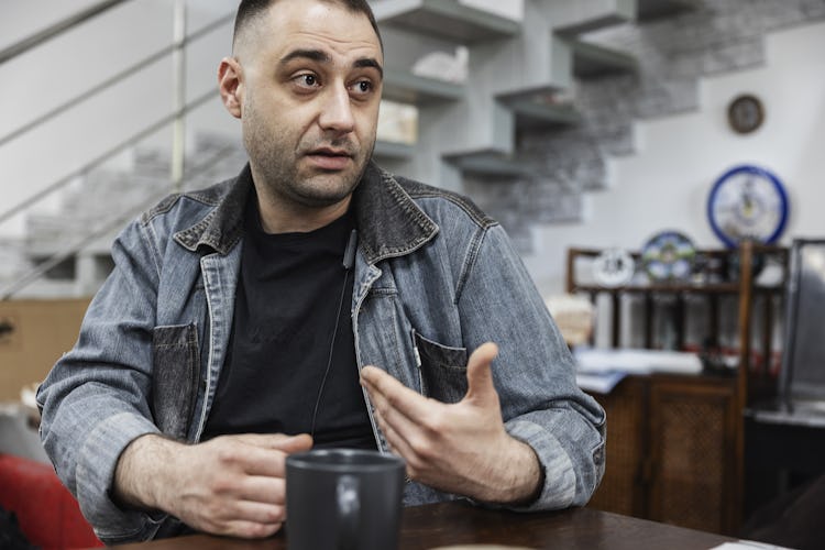 En mörkhårig man med skäggstubb, iklädd jeansjacka, sitter vid ett bord med en kaffekopp framför sig.