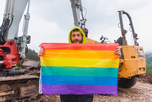 Skäggig man på byggarbetsplats håller i en regnbågsflagga.