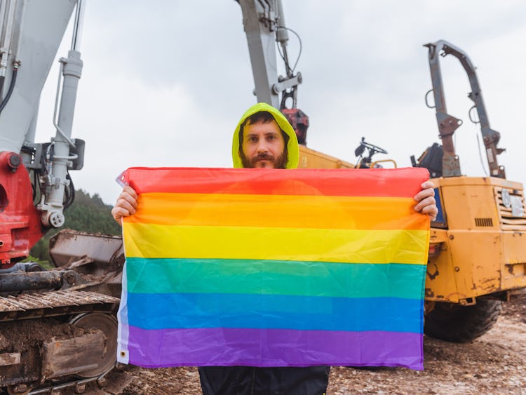 Skäggig man på byggarbetsplats håller i en regnbågsflagga.
