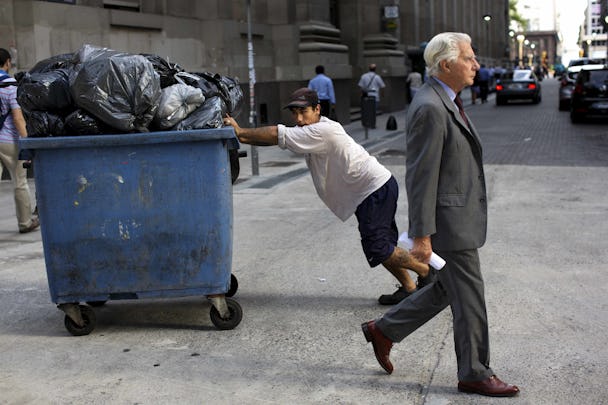 En så kallad lumpsamlare knuffar en container i Bunos Aires medan en man i kostym går förbi.
