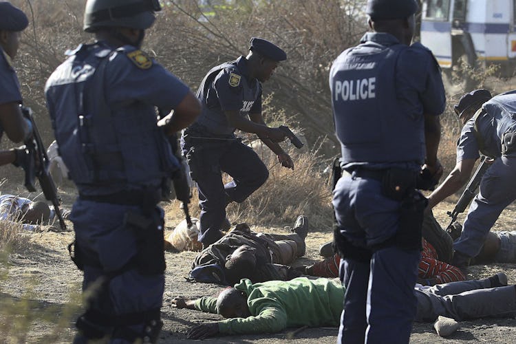 Sydafrikanska poliser med dragna vapen står runt skjutna gruvarbetare som ligger på marken.