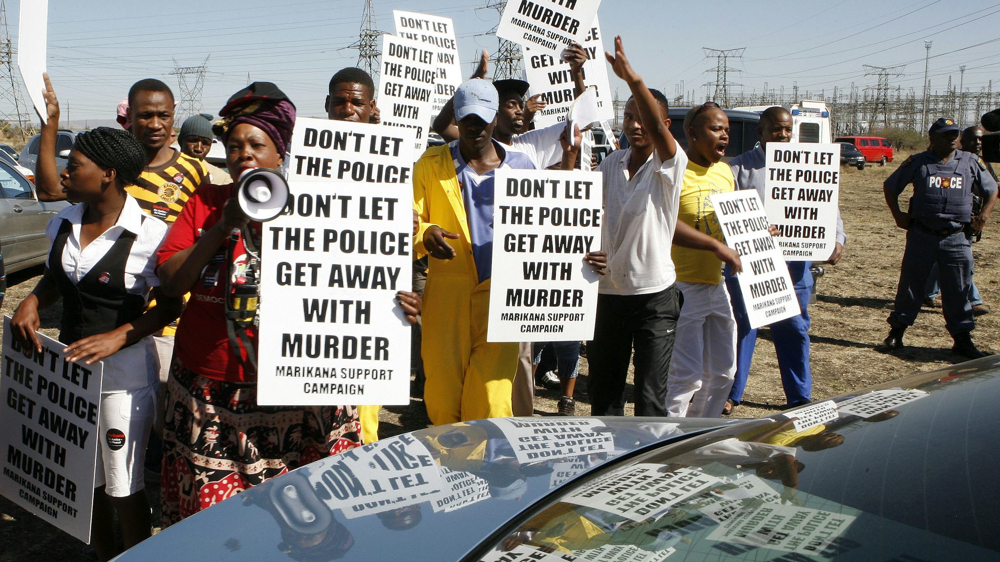 Sydafrikanska demonstranter med plakat står runt en bil. Texten på plakaten: "Låt inte polisen komma undan med mord."