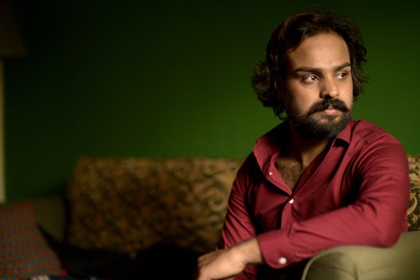 En pakistansk man i röd skjorta sitter i en soffa och blickar åt sidan. 