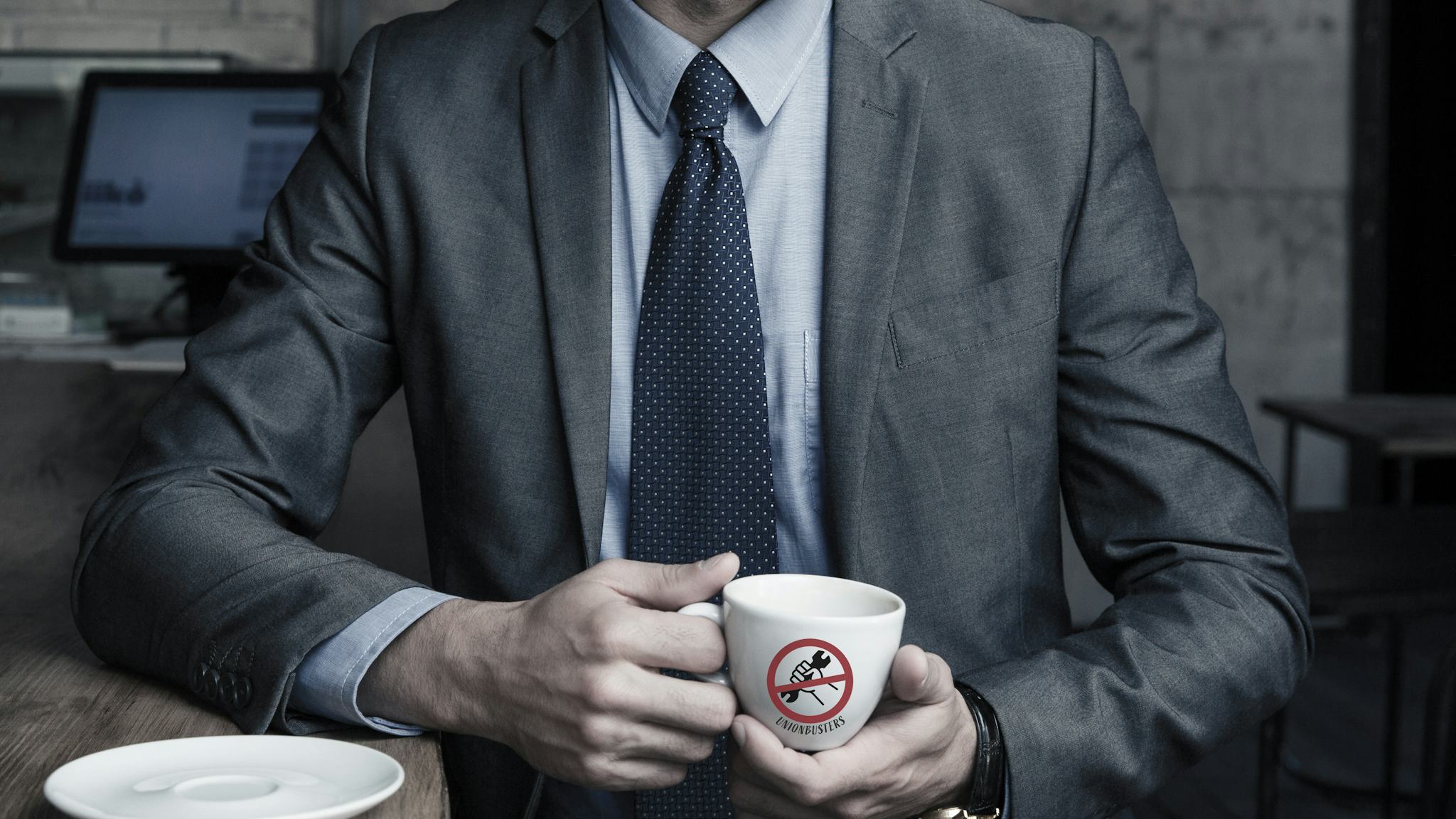 En man i grå kostym, vars ansikte inte syns, håller en kaffekopp med texten "Union busters".
