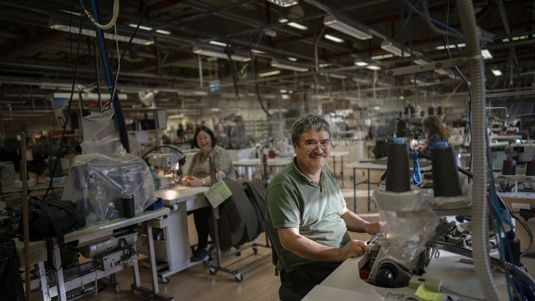 En medelålders man ler vid en symaskin, i bakgrunden skrattar en kollega.