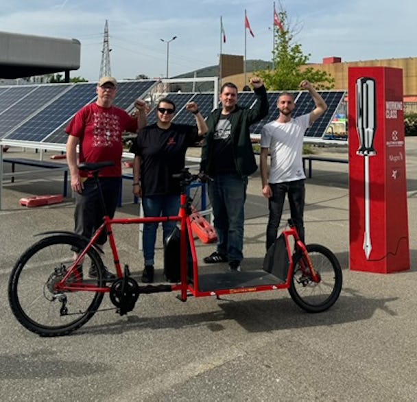 Fyra personer poserar med en stor röd tandemcykel vid en utomhusladdningsstation för elbilar.