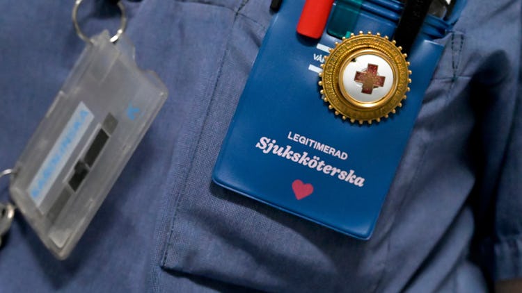 En närbild på en sjuksköterskas märke och pennor i en ficka, där identifikation och en röd hjärtsymbol syns, fäst på en blå uniform.