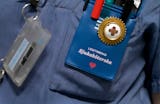 En närbild på en sjuksköterskas märke och pennor i en ficka, där identifikation och en röd hjärtsymbol syns, fäst på en blå uniform.