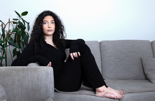 En kvinna med lockigt hår sitter i en grå soffa, klädd i svarta kläder, och tittar rakt in i kameran.