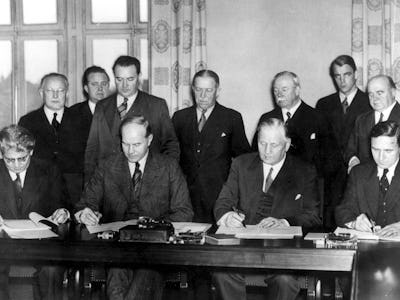 Nio kostymklädda män sitter vid ett långt bord och undertecknar dokument, med tre män som står bakom dem och observerar i ett historiskt svartvitt foto.