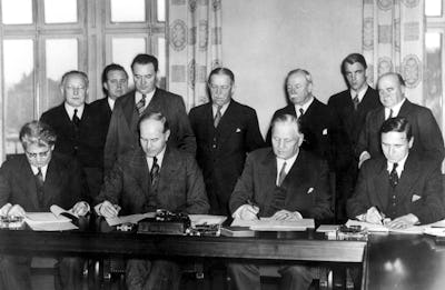 Nio kostymklädda män sitter vid ett långt bord och undertecknar dokument, med tre män som står bakom dem och observerar i ett historiskt svartvitt foto.