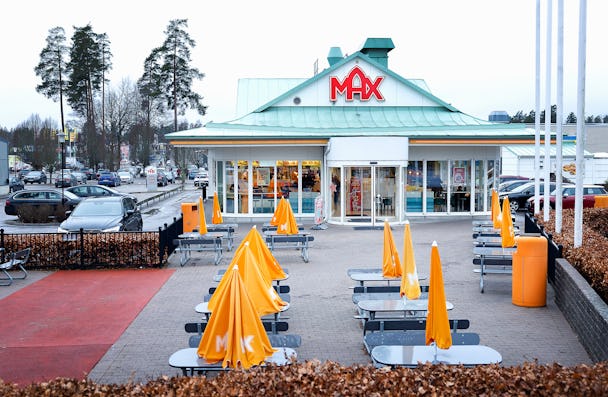 Alt text: Max snabbmatsrestaurang exteriör med uteservering, orange paraplyer, i en förortsmiljö med parkerade bilar och träd synliga i bakgrunden.