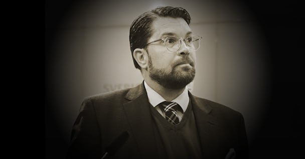 En man med skägg och glasögon, iförd kostym och randig slips, tittar åt sidan under en diskussion. Bilden är i sepia ton.