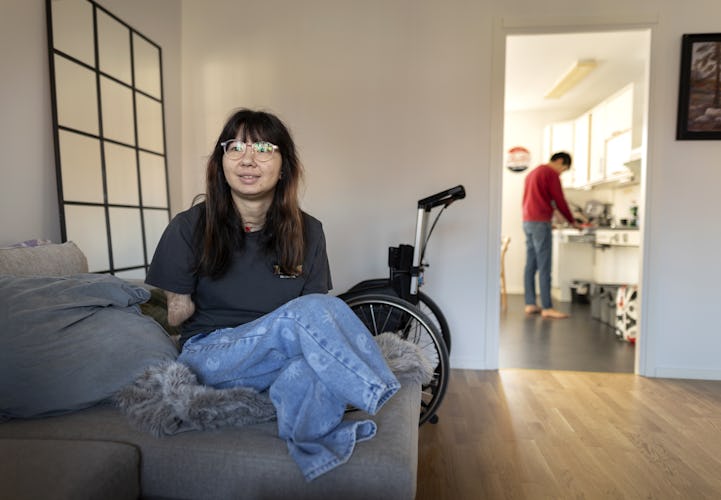 En kvinna i rullstol sitter på en soffa i ett vardagsrum, ler mot kameran, medan en annan person lagar mat i köket i bakgrunden.