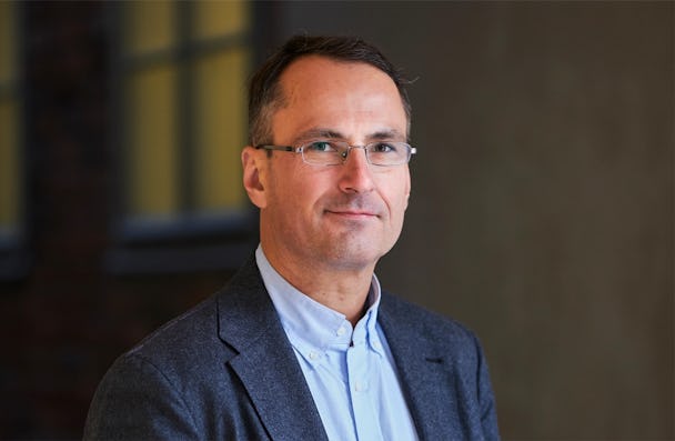 En medelålders man i glasögon, klädd i en grå kavaj och ljusblå skjorta, poserar med ett leende i kontorsmiljö.