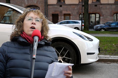 Kvinna i glasögon och blå jacka talar i röd mikrofon medan hon håller i anteckningar, vit bil och tegelbyggnader syns i bakgrunden.