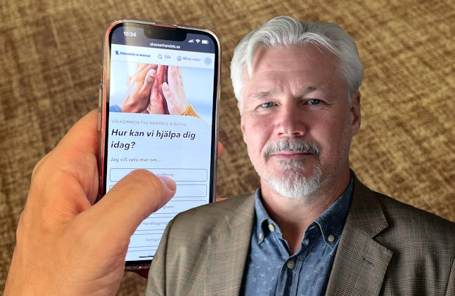 En hand håller en smartphone som visar en webbplats på svenska. Till höger står en medelålders man med grått hår och skägg, iklädd en blazer.