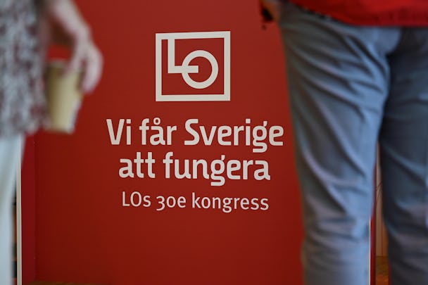 Två personer står framför en röd skylt med vit text som lyder "Vi får Sverige att fungera, LOs 30e kongress," under logotypen för Landsorganisationen i Sverige (LO).