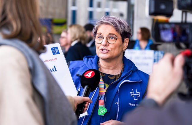 En kvinna med kort hår, glasögon och en blå jacka talar i en mikrofon märkt "4" i en utomhusmiljö. Det finns i människors bakgrunden. Hon håller ett dokument med synlig text i högre hand.