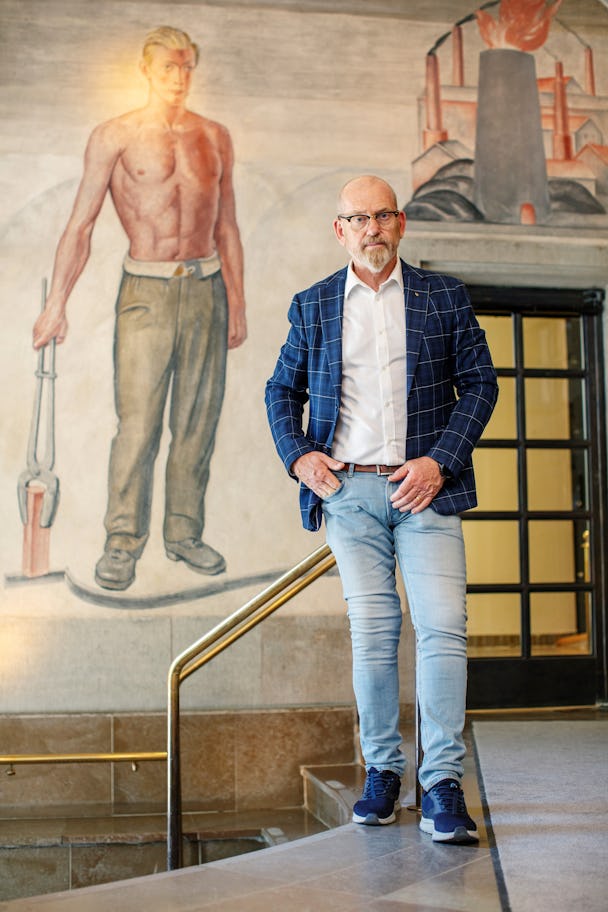 En man står på en trappa framför en väggmålning som visar en person utan skjorta som håller ett verktyg. Mannen har glasögon, blå kavaj och ljusa jeans.