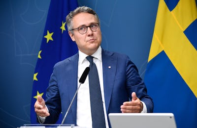En man i kostym och slips står framför en EU-flagga. Han talar vid ett podium med en mikrofon.