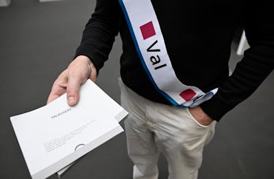 En person med ett band där det står "Val" håller i ett kuvert märkt "VALKUVERI" framför sin överkropp.