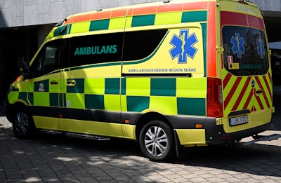 En gul och grön ambulans från Region Skåne står parkerad på ett asfalterat område, med ordet "Ambulans" och medicinska symboler synliga på dess sida och bakre del.