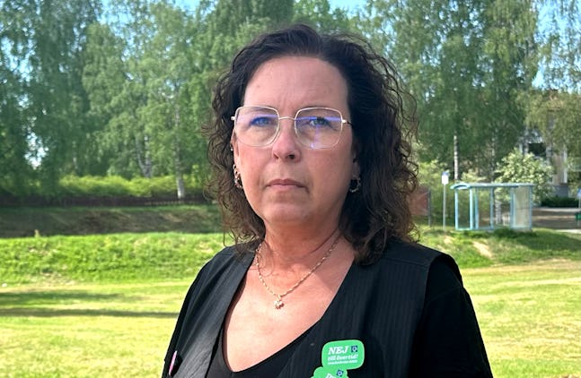 En kvinna med lockigt hår, glasögon, svart topp och en grön namnskylt står i ett gräsbevuxet område med träd i bakgrunden.