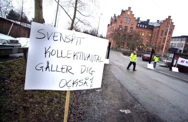 Protestbild med en skylt på svenska som lyder "Svenskt kollektivavtal gäller dig också!" samt två personer i varseljackor i bakgrunden.