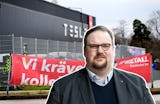 En man står framför en Tesla-anläggning. En röd banderoll med svensk text syns i bakgrunden, bredvid ett staket.