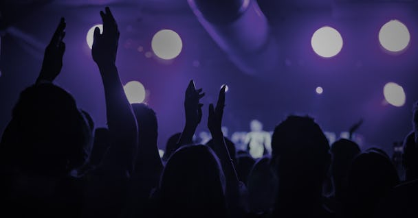 En folksamling med uppsträckta händer på en konsert under lila belysning, med upplysta cirkulära lampor i bakgrunden.