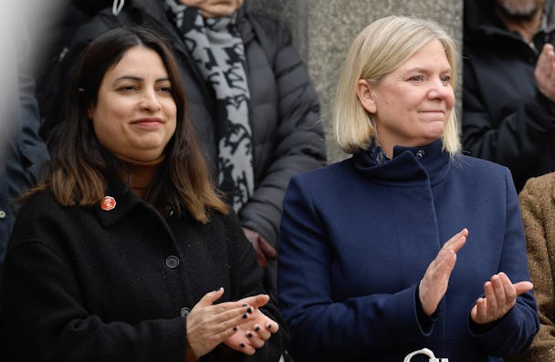Två kvinnor, en med mörkt hår och en med blont hår, står bredvid varandra och klappar. Kvinnan till vänster har på sig en mörk kappa medan kvinnan till höger bär en marinblå kappa.