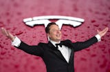 En man i smoking står med utsträckta armar framför en röd bakgrund med en Tesla-logga.