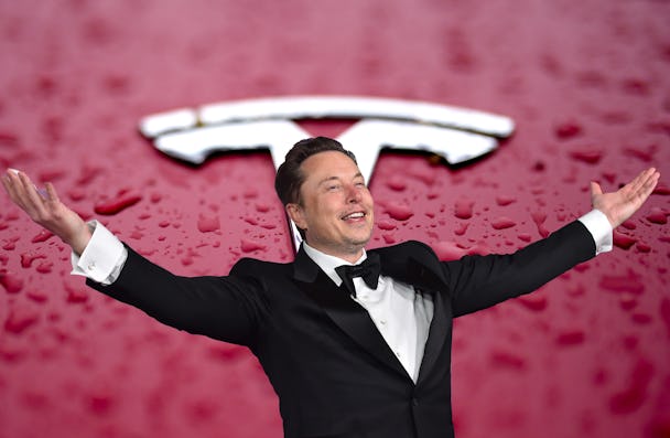 En man i smoking står med utsträckta armar framför en röd bakgrund med en Tesla-logga.